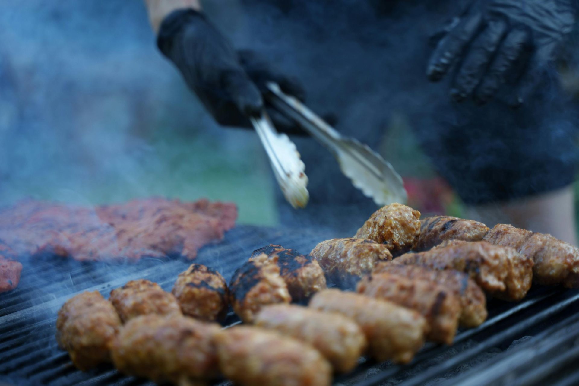 Nahaufnahme von brutzelndem Fleisch, das im Freien mit einer Grillzange gebraten wird, während Rauch aufsteigt.