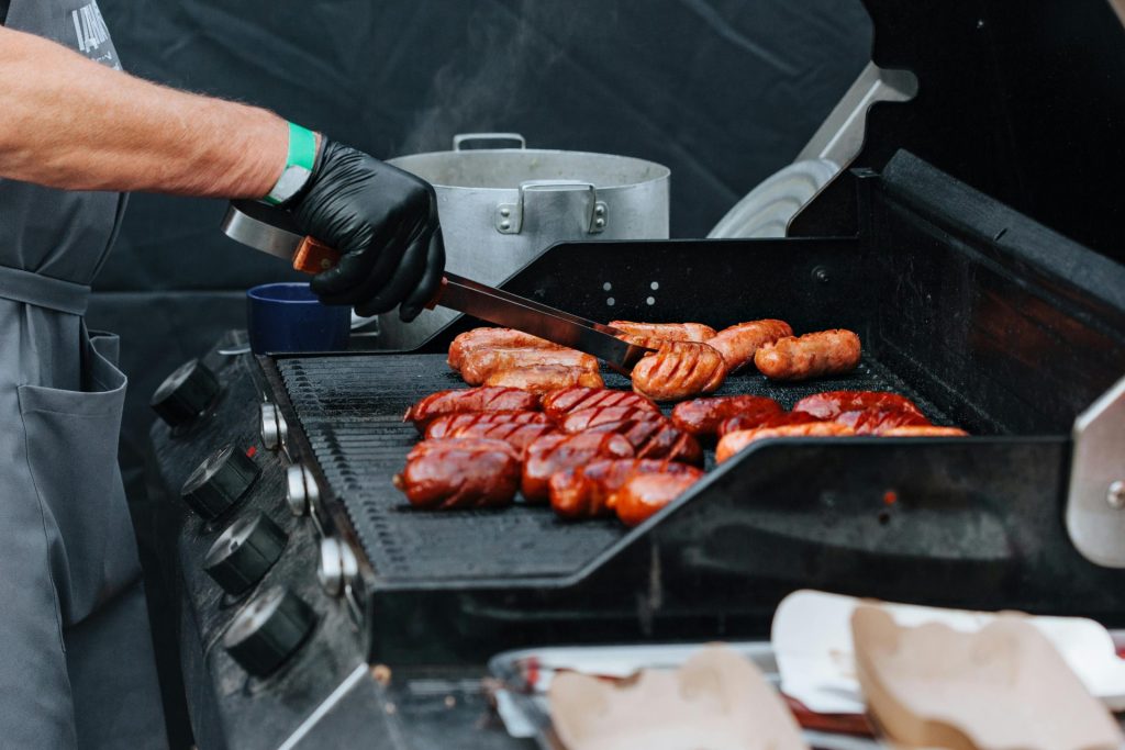 Nahaufnahme eines Kochs, der verschiedene Würste auf einem Außengrill zubereitet – perfekt für Sommerveranstaltungen.